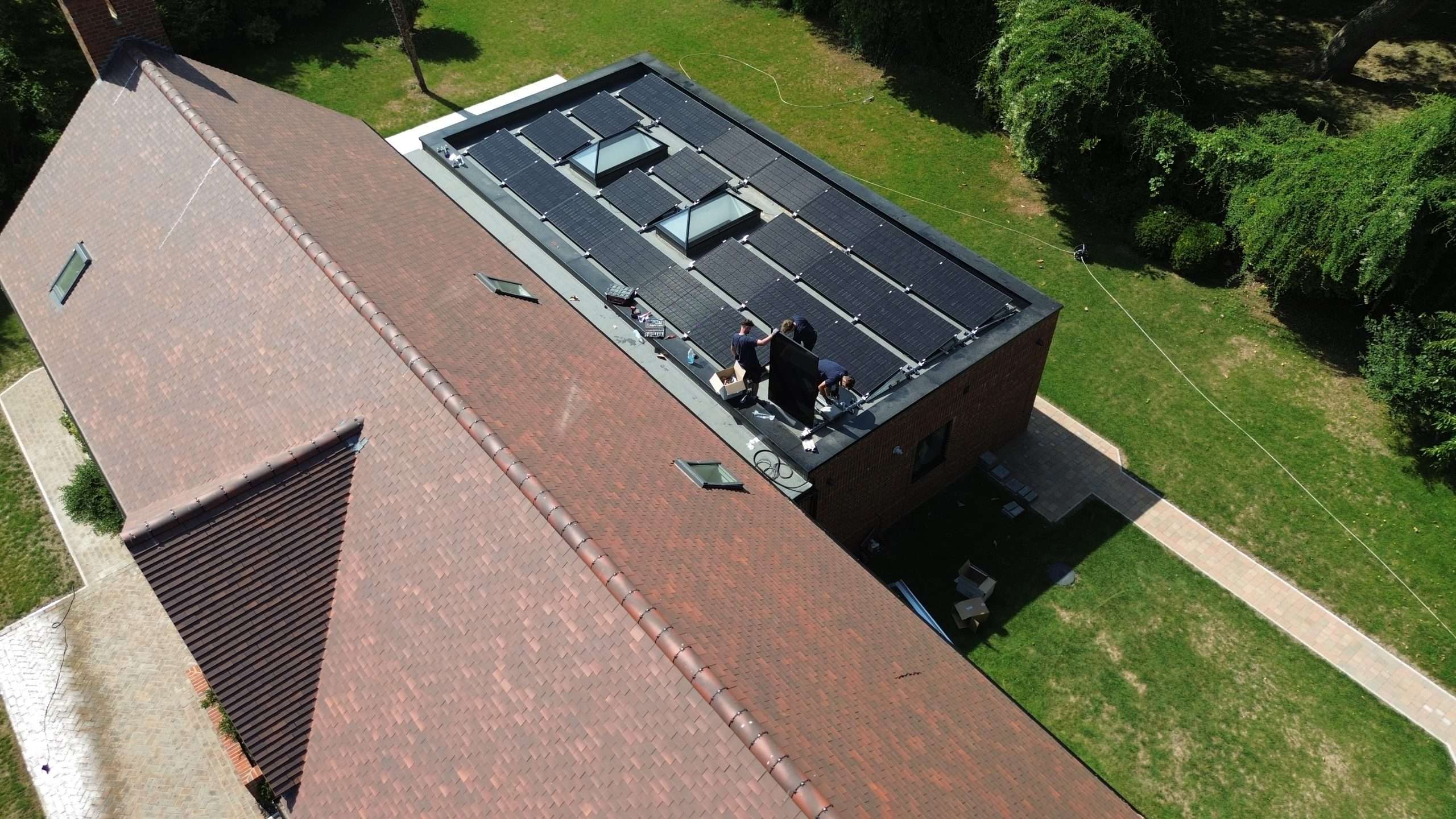Aerial view of installers fitting solar panels on the flat roof of a large residential property surrounded by gardens.